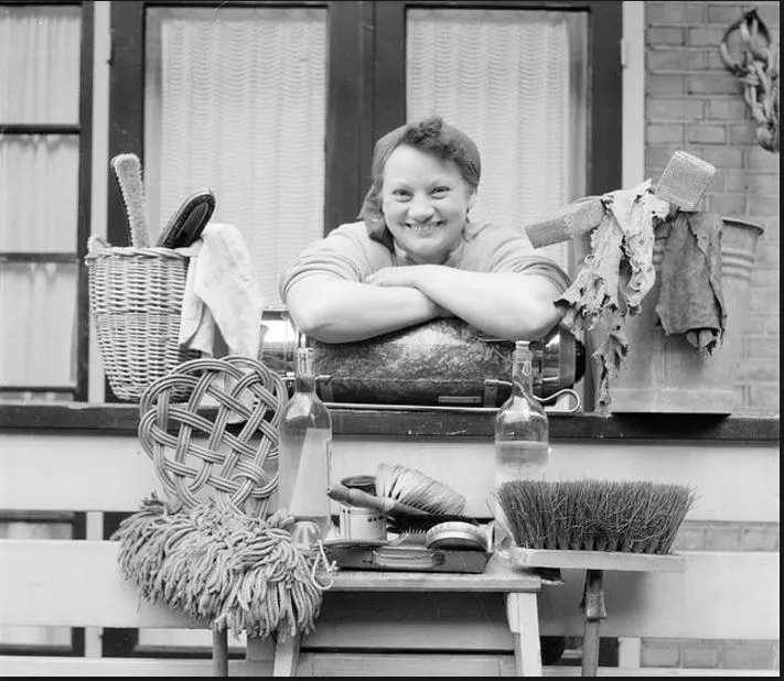 Huishoudelijke hulp kijkt lachend vanuit balkon met al haar schoonmaakspullen onder zich